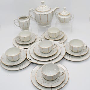 Könnte beinhalten: Ein weißes Porzellan-Teeservice bestehend aus einer Teekanne, einem Milchkännchen, einer Zuckerdose und sechs Tassen mit passenden Untertassen und Tellern. Jedes Teil hat ein gewelltes Paneel-Design mit dünnem Goldrand und vertikalen Rillen.