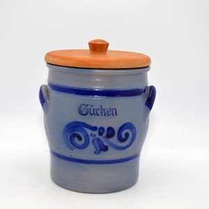 Può includere: Vaso in ceramica azzurra con coperchio in legno. Il vaso presenta dettagli e manici blu scuro, e la scritta "Gurken". Un motivo a spirale decorativo è dipinto sotto.