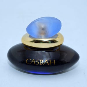 Pode incluir: Um frasco de perfume azul escuro com uma faixa dourada e uma tampa oval azul clara. A palavra "CASBAH" é impressa em ouro na frente do frasco. O frasco tem uma forma oval elegante.