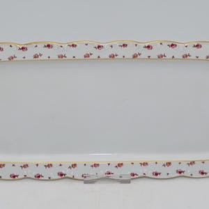 Puede incluir: Una bandeja rectangular de porcelana blanca con borde festoneado. El borde está decorado con un patrón repetido de pequeñas rosas rosas y detalles dorados. La bandeja está diseñada para servir alimentos.