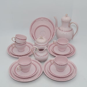 Op de afbeelding: Een complete roze porseleinen theeset met gouden accenten. De set bevat kopjes, schotels, borden, een theepot, een melkkan en een suikerpot. Het servies heeft een delicaat bloemenpatroon en geschulpte randen.
