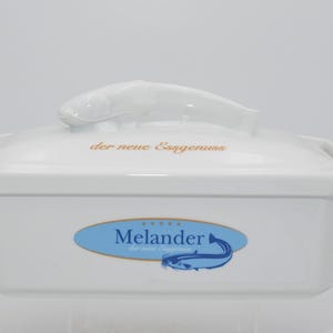 Può includere: Piatto da portata rettangolare in ceramica bianca con manico a forma di pesce sul coperchio. Il coperchio ha un testo dorato che recita "der neue Ergenuss". Il piatto presenta un logo ovale blu con il nome "Melander" e un'illustrazione di pesce.