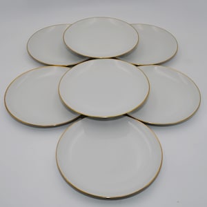 Op de afbeelding: Een set van zeven ronde witte borden gerangschikt in een groep. Elk bord heeft een glad, effen wit oppervlak en een dunne, metallic gouden rand langs de buitenkant. De borden zijn uniform in ontwerp, kleur en ronde vorm.