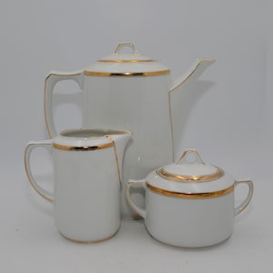Op de afbeelding: Een wit porseleinen theeservies met gouden rand. De set bevat een theepot, een melkkan en een suikerpot met deksel. De theepot heeft een hoog, hoekig ontwerp, terwijl de melkkan en suikerpot ronder zijn.