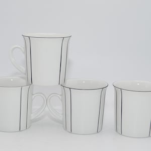 Op de afbeelding: Vier witte keramische mokken met een zwart lijnenontwerp. Elke mok heeft een handvat en een zwarte lijn die de vorm van de mok volgt. De mokken zijn gestapeld en gerangschikt op een witte achtergrond.