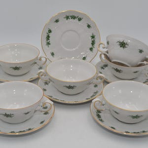 Può includere: Un set di tazze e piattini in porcellana bianca con bordi dorati e decorazioni floreali verdi. Il set comprende sei tazze, sei piattini e un piatto da portata. Le tazze hanno due manici e i piattini sono rotondi.