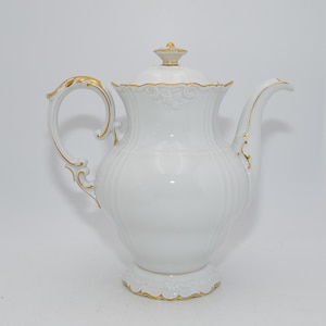 Könnte beinhalten: Eine weiße Porzellan-Kaffeekanne mit Deckel und gebogenem Ausguss. Der Griff und die Zierleisten sind goldfarben. Die Kanne hat ein geriffeltes Design und steht auf einem dekorativen Sockel. Ein goldfarbener Knauf krönt den Deckel.