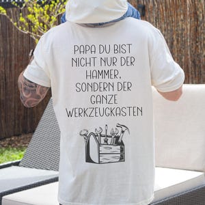 Könnte beinhalten: Weißes T-Shirt mit einem schwarzen Aufdruck, auf dem "Papa du bist nicht nur der Hammer, sondern der ganze Werkzeugkasten" steht. Der Aufdruck enthält auch eine Schwarzweißillustration einer Werkzeugkiste mit Werkzeugen.