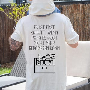 Könnte beinhalten: Cremefarbenes T-Shirt mit dem deutschen Text "ES IST ERST KAPUTT, WENN PAPA ES AUCH NICHT MEHR REPARIEREN KANN" und einer Werkzeugkasten-Grafik. Die Grafik enthält einen Schraubenschlüssel, einen Schraubendreher und einen Hammer. Das Shirt wird im Freien getragen.
