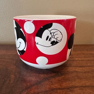 May include: A red and white ceramic mug featuring Mickey Mouse. The mug has a red background with white polka dots and black and white images of Mickey Mouse. The interior of the mug is white.