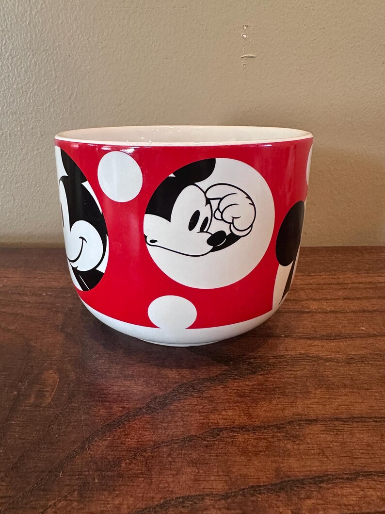 May include: A red and white ceramic mug featuring Mickey Mouse. The mug has a red background with white polka dots and black and white images of Mickey Mouse. The interior of the mug is white.