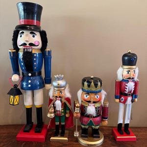 May include: Four decorative wooden nutcrackers in various sizes and designs. The nutcrackers feature a variety of colors, including blue, red, black, and gold. One nutcracker holds a lantern. The nutcrackers are standing on a wooden surface.