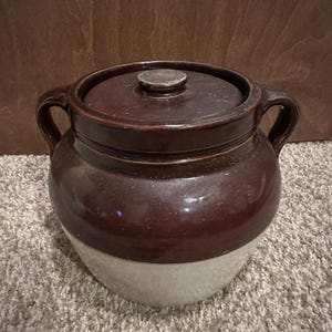 Könnte beinhalten: Ein antiker Steinguttopf mit Deckel in zweifarbigem Design. Der obere Teil ist dunkelbraun, der untere Teil cremefarben. Er hat Griffe und einen Knopf auf dem Deckel. Der Topf steht auf einem neutralen Teppich.