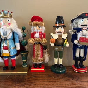 Könnte beinhalten: Vier dekorative Nussknacker aus Holz in verschiedenen Outfits. Einer trägt eine Krone und einen blauen Mantel, ein anderer einen Weihnachtsmann-Anzug, ein dritter ein Pilger-Outfit und der letzte einen blauen Mantel und Hut. Jeder hält einen kleinen Gegenstand.