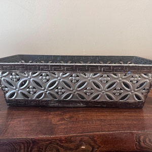Peut inclure: Panier rectangulaire en métal avec une finition vieillie. Le panier présente un motif décoratif de cercles et de fleurs, avec une bordure marron foncé et un intérieur gris clair. Le panier est vide et posé sur une surface en bois.
