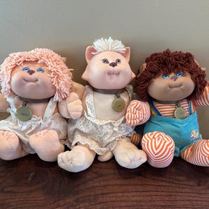 May include: Three plush dolls with round faces and soft bodies. The left doll has pink yarn hair and a white floral romper. The center doll is a cat-like figure with white hair and a white dotted dress. The right doll has brown hair and blue overalls.