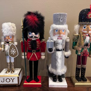 May include: Four decorative wooden nutcrackers in various outfits. One wears a black and red fur hat and red jacket. Another has a white fur hat and holds a shield. The third wears a gray coat and hat. The last one has a black sequined hat and green jacket.
