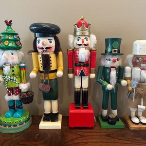 May include: Five wooden nutcrackers in various designs. One holds a staff with a birdhouse and Christmas tree hat. Others wear hats and uniforms, including a chef, a king, and a leprechaun. They stand on individual bases, with a variety of colours.