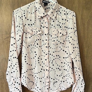 Può includere: Camicia rosa chiaro con bottoni e un motivo a cuori neri. La camicia a maniche lunghe ha due tasche sul petto e un colletto. Il nome del marchio "wet seal" è visibile sul colletto.