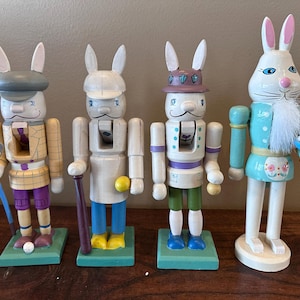Puede incluir: Cuatro cascanueces de conejito de Pascua de madera. Cada conejito está pintado con diferentes atuendos y accesorios, incluidos palos de golf, bates de béisbol y una canasta de huevos. Los conejitos están sobre bases verdes.