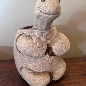 May include: A beige, textured ceramic planter shaped like a seated frog. The frog has a smooth, smiling face with dark brown eyes and holds a sculpted flower against its chest. The planter features a hollow opening at the top for plants.