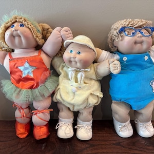May include: Three Cabbage Patch Kids dolls standing side by side. The left doll has blonde hair, an orange outfit with a silver star, and orange shoes. The center doll wears a yellow hat and outfit. The right doll has blonde hair, blue glasses, and a blue romper.