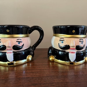 Pode incluir: Duas canecas de cerâmica em forma de quebra-nozes. Cada caneca tem um chapéu preto, um rosto rosa, um bigode preto e uma boca vermelha. As canecas têm alças pretas e detalhes dourados.
