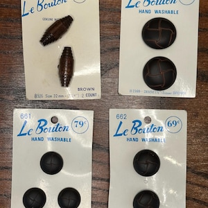 Peut inclure: Quatre cartes de boutons vintage. Les cartes présentent la marque "Le Bouton" en écriture bleue. Les boutons sont marron et sont disponibles en différentes tailles, dont 32 mm, 28 mm, 22 mm et 19 mm.