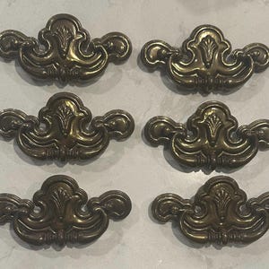 May include: Six antique brass drawer pulls with an ornate, symmetrical design. Each pull features a central decorative element flanked by curved handles. The brass has a warm, aged patina, suggesting vintage or retro style. These pulls are suitable for furniture restoration.