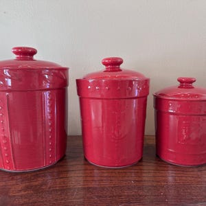 Op de afbeelding: Drie rode keramische keukenpotten met deksels. De potten hebben verschillende maten, de grootste links en de kleinste rechts. De potten hebben een textuur en een ronde knop op het deksel.