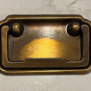May include: A brass cabinet pull with a rectangular shape and a rounded edge. The pull has a recessed centre and two small, round knobs on either side.