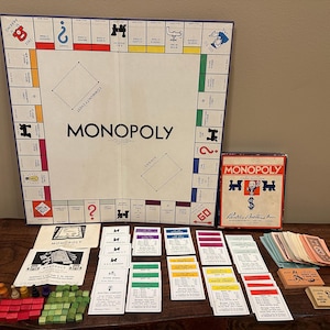 Puede incluir: Juego de mesa Monopoly con tablero, cartas, dinero de juguete y fichas. El tablero es blanco con casillas de colores y la palabra "MONOPOLY". La caja es roja y blanca.