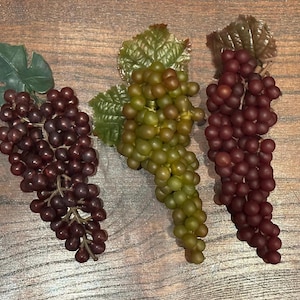 Puede incluir: Tres racimos de uvas artificiales dispuestos en fila. Dos racimos son de color morado oscuro y uno central es de color verde claro. Cada racimo presenta frutos pequeños, redondos y esféricos unidos a un tallo central con una hoja verde.