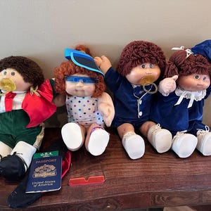 May include: Four Cabbage Patch Kids dolls with yarn hair, wearing various outfits including a green jacket, a polka dot swimsuit with blue visor, a blue suit, and a blue dress. A U.S.A. passport and a pair of red-rimmed glasses are placed in front.