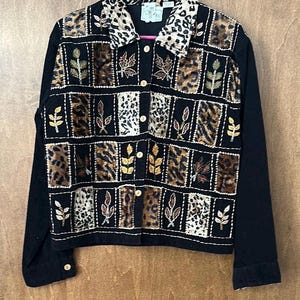 Puede incluir: Chaqueta negra con mangas largas y cierre de botones. La chaqueta presenta un cuello con estampado de leopardo y un diseño de patchwork en la parte delantera con cuadrados de hojas y estampado de leopardo. Detalles de hojas bordadas en oro y marrón.