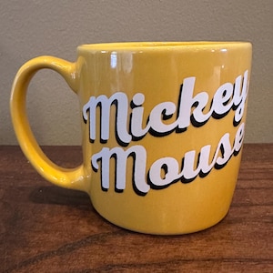 May include: A yellow ceramic mug with the words "Mickey Mouse" in white script with a black outline. The mug has a curved handle and is set on a wooden surface.