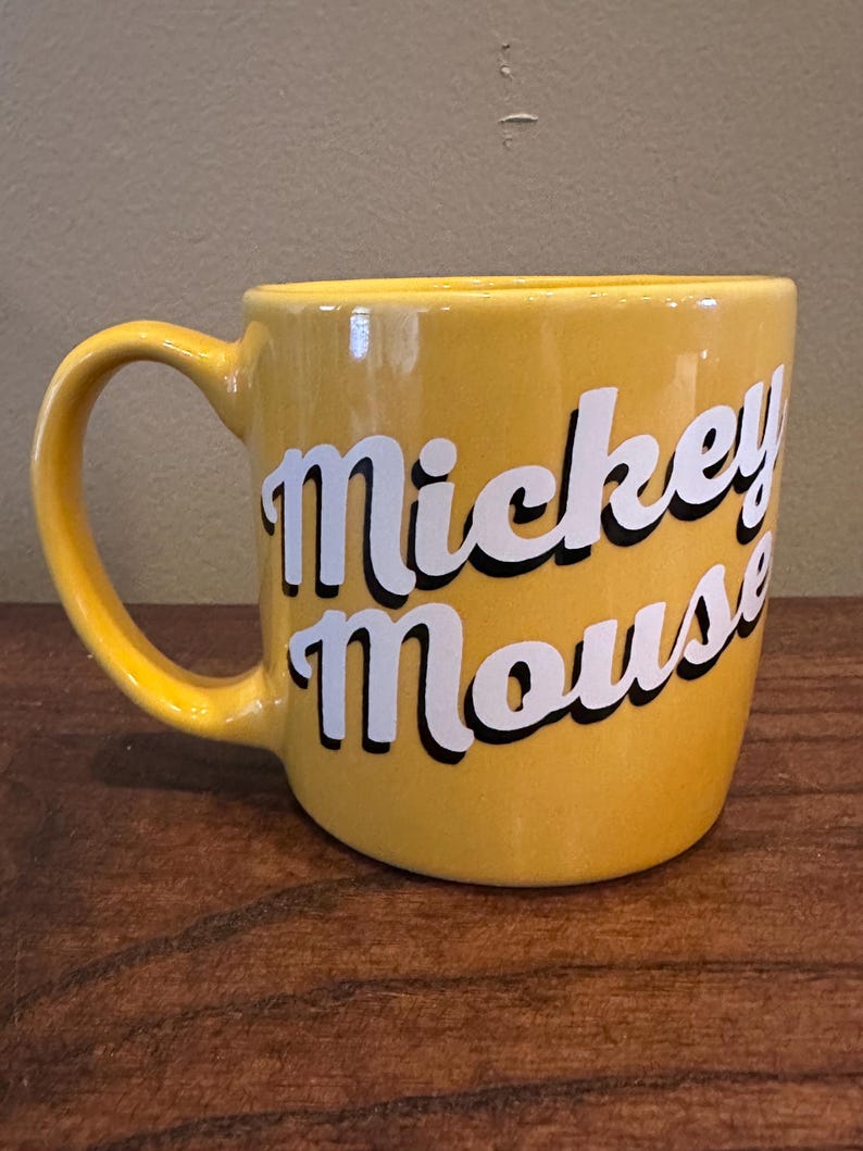 May include: A yellow ceramic mug with the words "Mickey Mouse" in white script with a black outline. The mug has a curved handle and is set on a wooden surface.