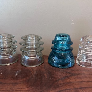Pode incluir: Quatro isoladores elétricos de vidro dispostos em fila. Dois são transparentes com cristas em camadas, um é de um azul-petróleo vibrante com o texto em relevo MADE IN USA, e o último é transparente com nervuras horizontais na base.