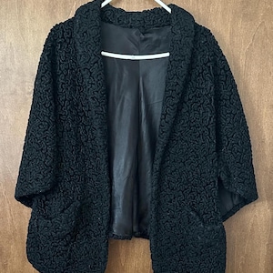 Puede incluir: Chaqueta negra texturizada con cuello chal y dos bolsillos delanteros grandes. La chaqueta está colgada de una percha blanca sobre un fondo de madera. El forro interior de la chaqueta es negro.