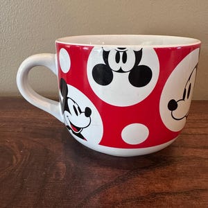 May include: White ceramic mug with a red polka dot pattern and Mickey Mouse character faces. The mug has a white handle and a wide, rounded shape. The design features Mickey's face in black and white.