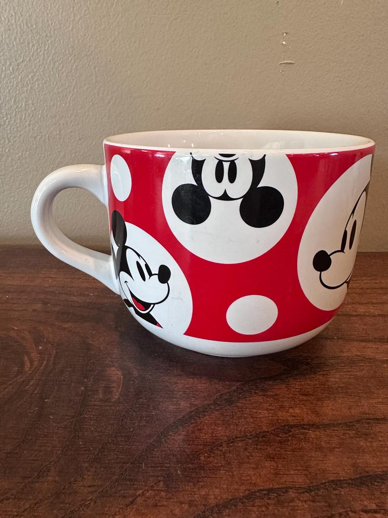 May include: White ceramic mug with a red polka dot pattern and Mickey Mouse character faces. The mug has a white handle and a wide, rounded shape. The design features Mickey's face in black and white.