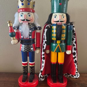 Puede incluir: Dos cascanueces decorativos de madera. Uno lleva una corona azul y dorada, un uniforme rojo y azul y sostiene un cetro. El otro lleva un sombrero verde con ribete dorado, una capa roja con lunares blancos y un uniforme verde y amarillo.