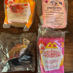 May include: Four sealed McDonald's Happy Meal toys in plastic packaging. One orange package features a Ronald McDonald graphic and the text "PATO LUCAS". Another pink package reads "CONGRATULATIONS On Your New Arrival!" The other two packages are pink and clear.