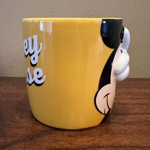 May include: Yellow ceramic mug featuring a 3D Mickey Mouse design on the side. The mug has the words "Mickey Mouse" in white script with a black outline. The mug is on a wooden surface.