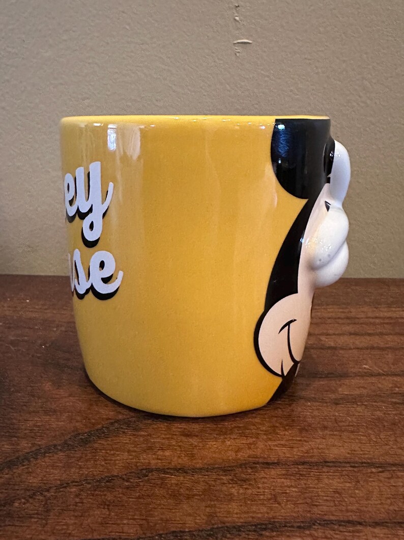 May include: Yellow ceramic mug featuring a 3D Mickey Mouse design on the side. The mug has the words "Mickey Mouse" in white script with a black outline. The mug is on a wooden surface.
