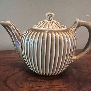 Op de afbeelding: Een keramische theepot met een glanzende, lichtbruine afwerking en een verticale ribbelstructuur. De theepot heeft een gebogen tuit, een bijpassend deksel met een kleine knopgreep en een stevig, gebogen handvat aan de zijkant.