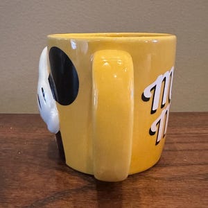 May include: Yellow ceramic mug with a handle. The mug features a white gloved hand with a black circle and the word "Mickey" in white script. The mug is sitting on a wooden surface.