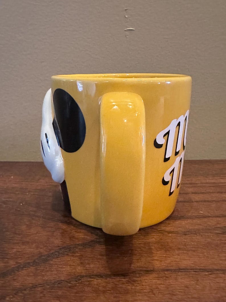 May include: Yellow ceramic mug with a handle. The mug features a white gloved hand with a black circle and the word "Mickey" in white script. The mug is sitting on a wooden surface.