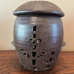 Pode incluir: Um pote de cerâmica castanho-escuro com tampa a condizer, apresentando uma superfície texturizada e desenhos recortados de caules e folhas de bambu. O recipiente tem um corpo arredondado, uma borda ligeiramente alargada e detalhes.