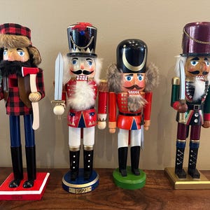 Könnte beinhalten: Vier Holz-Nussknacker in verschiedenen Designs. Einer trägt einen karierten Hut und Mantel und hält eine Axt und einen kleinen Baum. Andere haben Uniformen und Hüte im Militärstil und halten Schwerter.